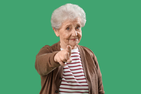 Senior woman pointing at viewer on green background. Accusation concept