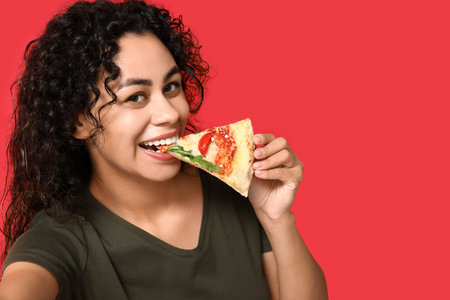 Beautiful young happy African-American woman with slice of delicious pizza taking selfie on red...