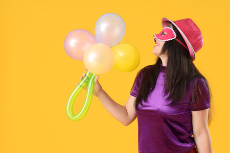 Pretty young woman in carnival mask with bouquet made from balloons on yellow backgroundの写真素材