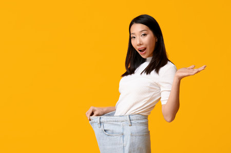Beautiful young shocked Asian woman in loose jeans on yellow background