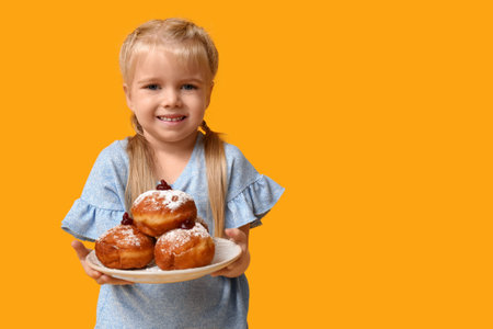 Cute happy little girl holding plate with tasty donuts on yellow background. Hanukkah celebrationの写真素材