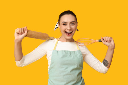 Portrait of happy young housewife in apron with whiskey and rolling pin on yellow backgroundの写真素材