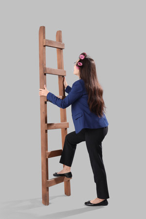 Funny Asian businesswoman with hair curlers climbing up ladder on gray backgroundの写真素材