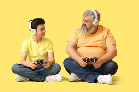 Happy grandfather with his cute grandson in headphones playing video games on yellow background