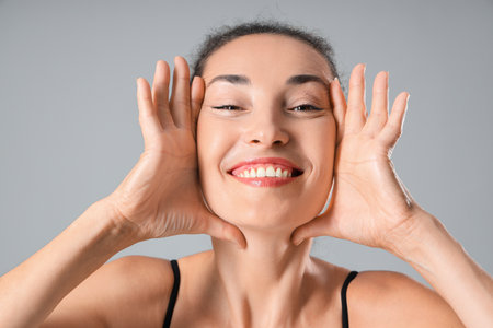 Beautiful mature woman giving herself face massage on gray backgroundの写真素材