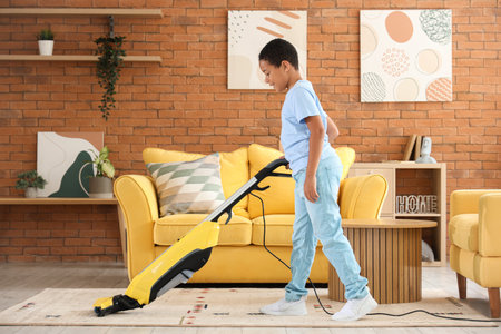 Cute African-American boy with vacuum cleaner cleaning carpet in living roomの写真素材