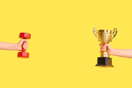 Female hands with gold cup and dumbbell on yellow backgroundの写真素材