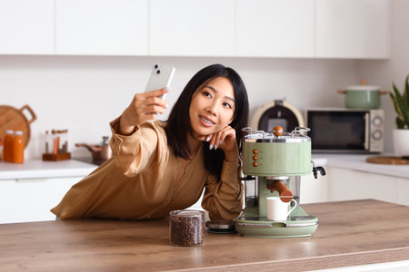 Beautiful Asian woman with coffee machine taking selfie in kitchenの写真素材