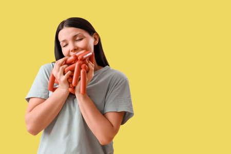 Young woman with sausages on yellow background. Overeating conceptの写真素材