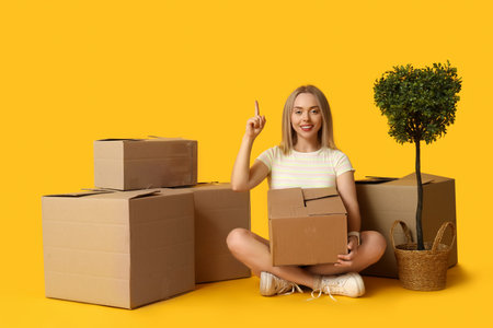Happy young woman with cardboard boxes and houseplant pointing at something on yellow background....