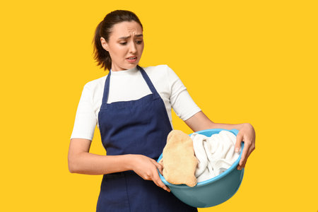 Portrait of tired young housewife in apron with dirty laundry on yellow backgroundの写真素材