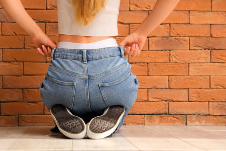 Young woman in stylish jeans pants sitting near orange brick wall, back viewの写真素材