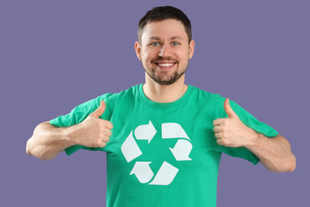Happy young man in t-shirt with recycling logo showing thumbs-up on lilac background. Ecology conceptの写真素材