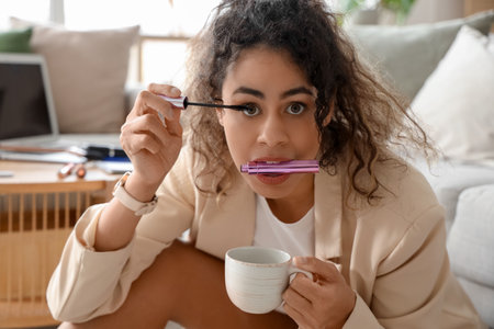 Hurrying African-American businesswoman with coffee cup applying mascara at home, closeupの写真素材