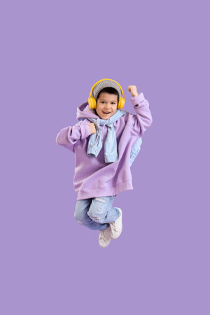 Cute little Asian boy in adult clothes with headphones jumping on lilac background