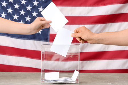 Voting woman and man near ballot box on table against flag of USAの写真素材