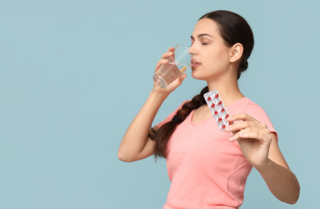Pretty young woman holding blister pack of vitamin A capsules and glass of water on blue backgroundの写真素材