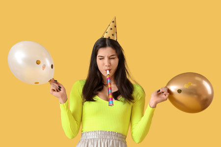 Drunk young woman with whistle and balloons after Birthday party on yellow backgroundの写真素材