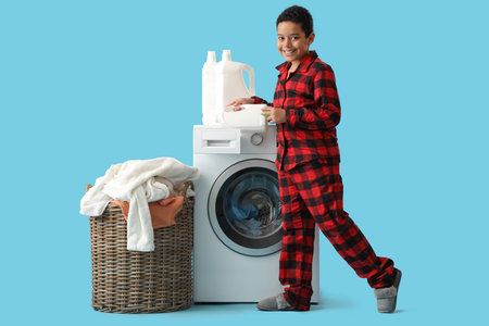 Smiling African-American boy pouring laundry detergent into washing machine near basket with dirty clothes on blue backgroundの写真素材