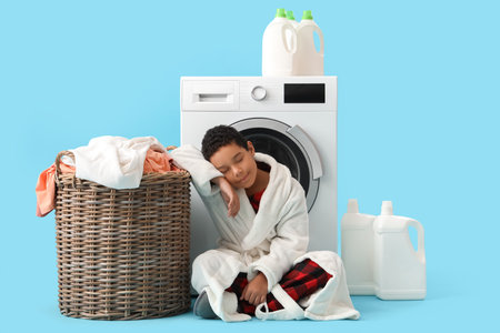 Little African-American boy sleeping near basket with dirty clothes and washing machine against blue backgroundの写真素材