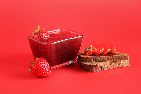Bowl with sweet strawberry jam and bread on red background