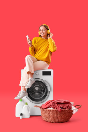 Pretty young woman sitting on washing machine and listening to music against red backgroundの写真素材