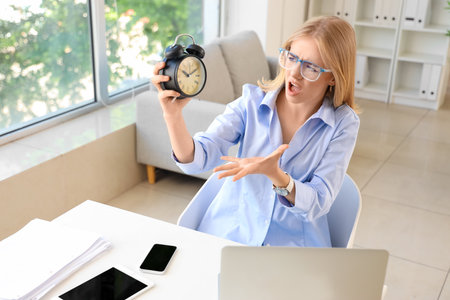 Stressed young businesswoman with alarm clock working under deadline in officeの写真素材