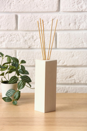 Bottle of reed diffuser and houseplant on table near light brick wall in room, closeup