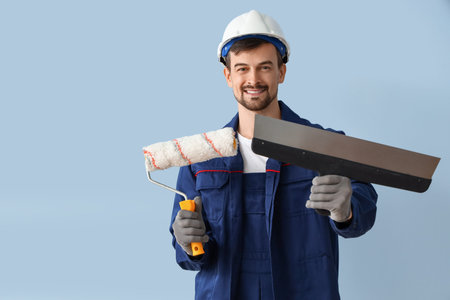 Male decorator with putty knife and paint roller on blue background