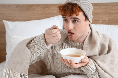 Ill young man eating soup in bedroomの写真素材
