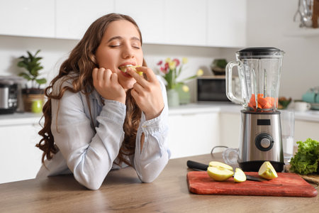 Beautiful young woman eating apple while making smoothie in kitchenの写真素材