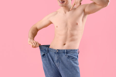 Handsome happy young sporty man pointing at loose jeans on pink background, closeup. Weight loss conceptの写真素材