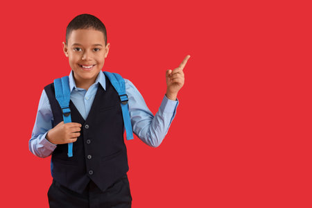 Happy little African-American schoolboy with backpack pointing at something on red backgroundの写真素材