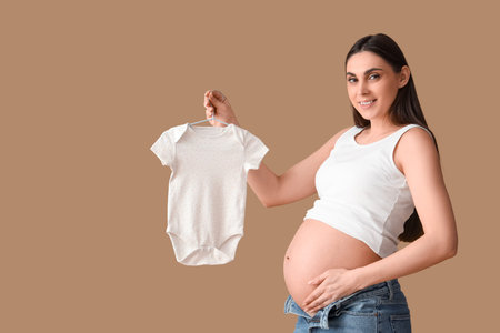 Young pregnant woman with baby bodysuit on brown background