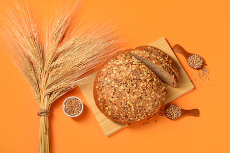 Wooden board with sliced loaf of bread, seeds and wheat spikelets on orange background