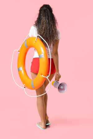 Beautiful young African-American female lifeguard with ring buoy and megaphone on pink background, back viewの写真素材