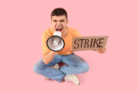 Protesting young man shouting into megaphone and holding placard with the word STRIKE on pink...