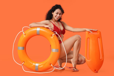 Beautiful young happy African-American female lifeguard with ring buoy and board on orange backgroundの写真素材