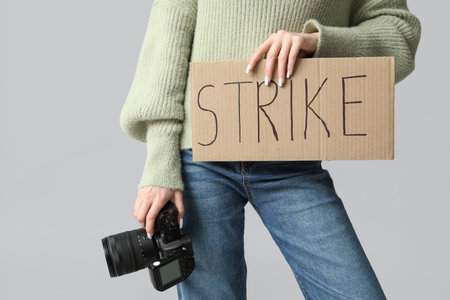 Protesting female photographer holding placard with text STRIKE and photo camera on gray background
