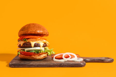 Board with tasty burger, onion rings and tomatoes on orange backgroundの写真素材
