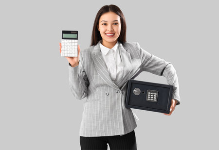 Beautiful young happy Asian woman with safe box and calculator on gray backgroundの写真素材
