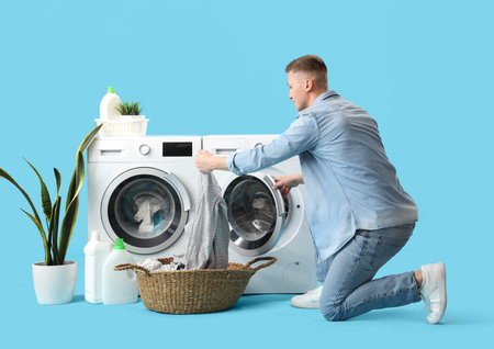 Handsome young man putting laundry into washing machine on blue backgroundの写真素材