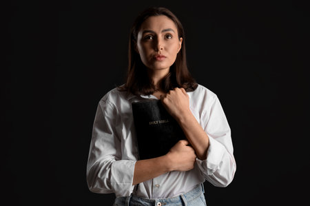 Young woman with Holy Bible praying on black background