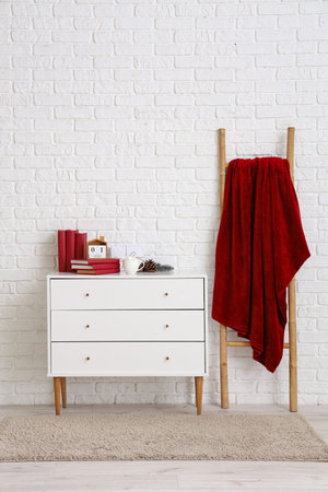 Chest of drawers with books and warm blanket on ladder near white brick wall