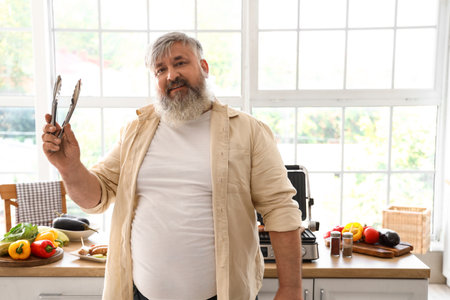 Mature man with tong cooking tasty sausages and vegetables on modern electric grill at table in kitchenの写真素材