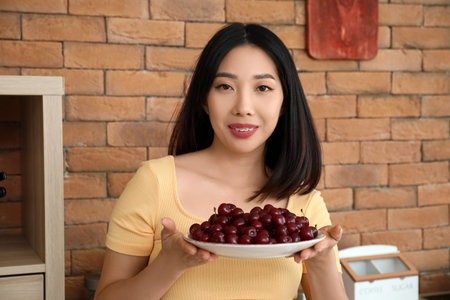 Happy beautiful young Asian woman with bowl of ripe cherries in kitchen