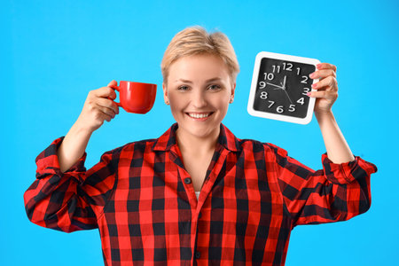 Young woman in pajamas with cup of coffee and clock on blue background
