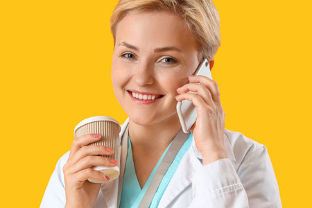 Female medical intern with cup of coffee talking by mobile phone on yellow background, closeupの写真素材