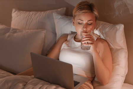 Young woman with laptop drinking water in bed at nightの写真素材
