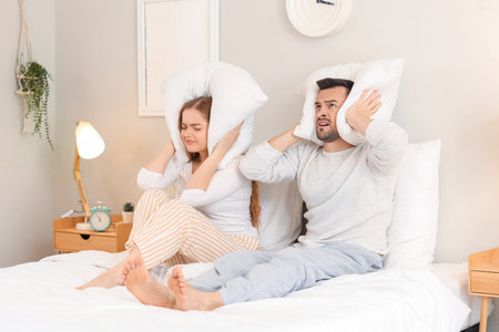 Young couple with pillows suffering from loud neighbors in bedroom